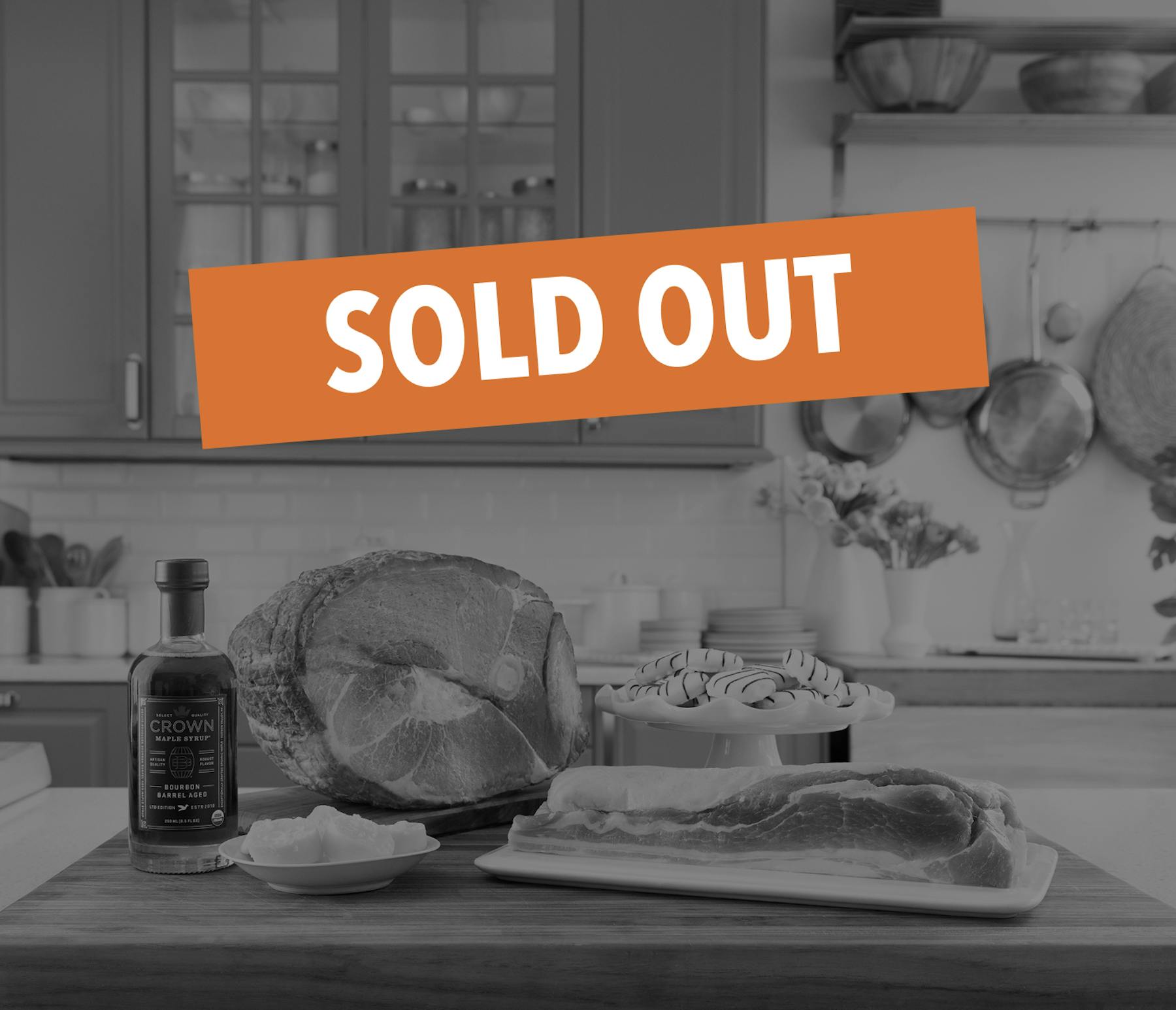 Bone-in heritage ham on wooden cutting board in bright kitchen, accompanied by bacon slices, Crown Maple Bourbon Syrup, butter, and white chocolate treats. Gray cabinets, subway tile, cookware, and spring flowers create an elegant cooking setting.